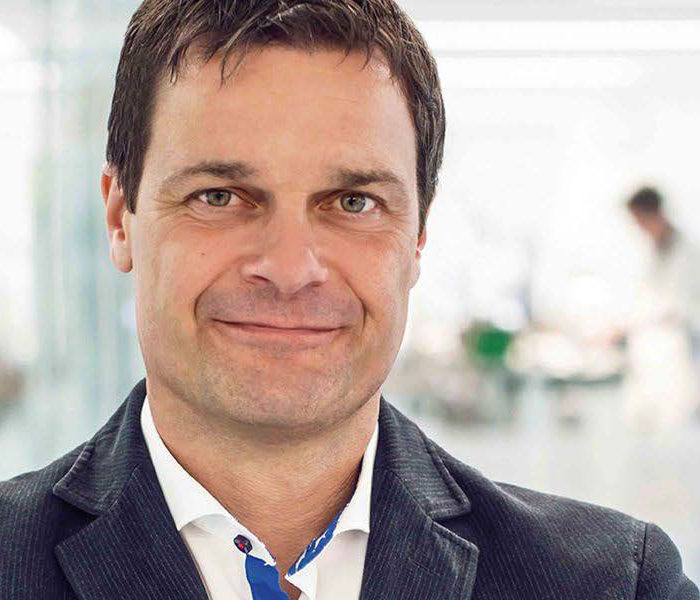 Porträt von Thilo Mühle im dunklen Anzug, im Hintergrund ist die Produktionsabteilung in der Manufaktur verschwommen zu sehen.