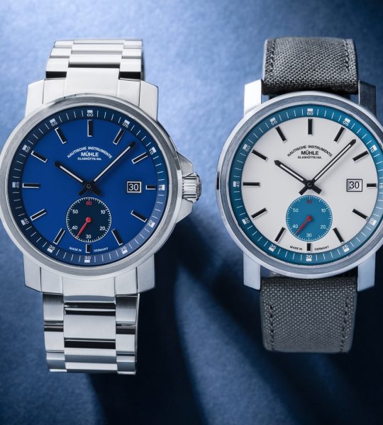 The 29er Big Small Seconds with blue dial and metal bracelet and the 29er Big Small Seconds with white-petroleum dial and grey strap lie side by side on a blue background
