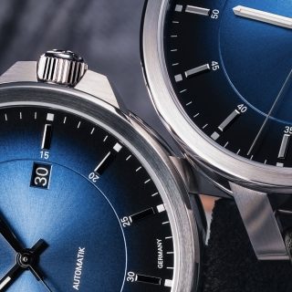 Bildausschnitt von zwei Armbanduhren (29er Casual Küstenblau) liegen nebeneinander auf einer grauen Oberfläche. Fokus liegt dabei auf den silbernen Gehäusen.