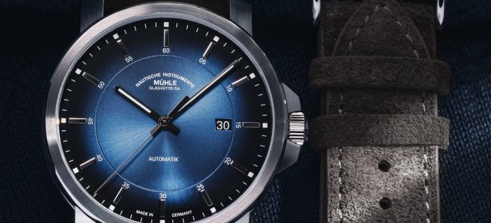 Armbanduhr 29er Casual Küstenblau am braunen Lederband liegt auf einem dunklen Untergrund, daneben ein graues Velourslederband.