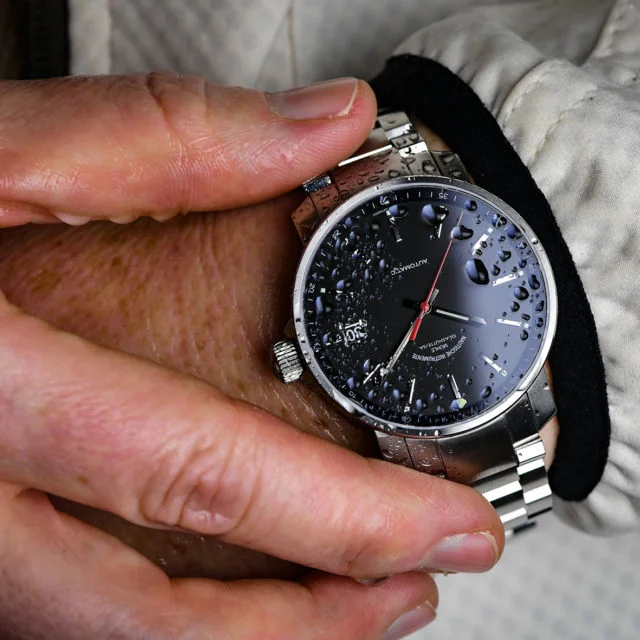 Wristshot der 29er mit schwarzem Zifferblatt am Metallband. Eine Hand greift nach der Uhr.