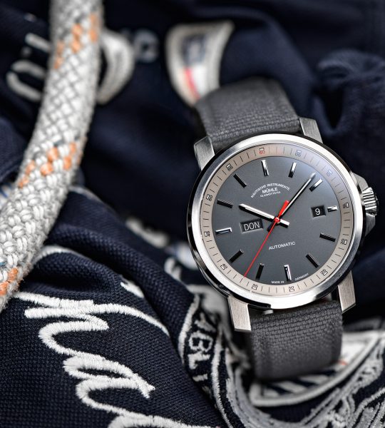 Herrenarmbanduhr 29er Big Tag/Datum mit grauem Zifferblatt am grauen Canvasband steht leicht seitlich auf blau-weißen Stoffen und Seilen.