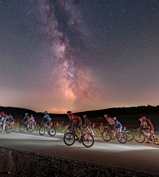 Nachtaufnahme von mehreren Rennradfahrern mit lila-farbenem Nachthimmel im Hintergrund.