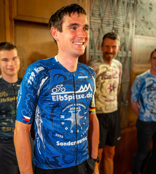 Ein Mann mit blauem Trikot von Mühle-Glashütte lächelt in die Kamera. Dahinter drei weitere Männer mit bunten Trikots.