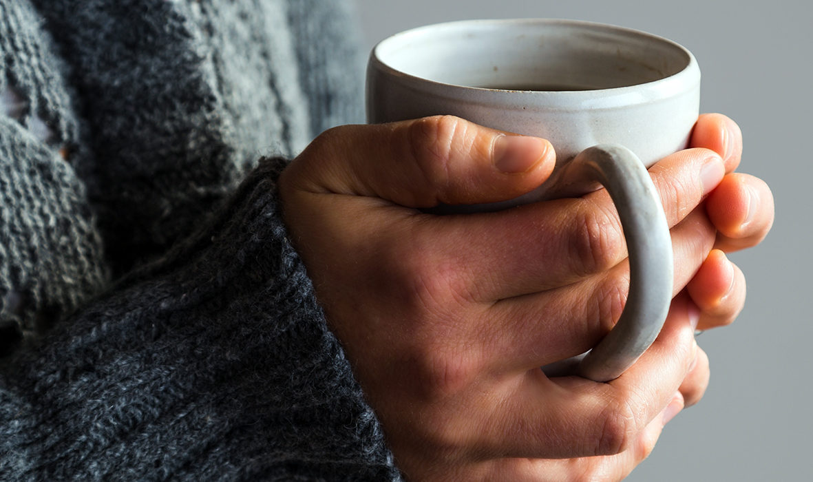Hände einer Person mit grauem Pullover, die eine weiße Kaffeetasse halten.