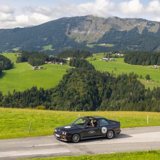 Ein schwarzes Auto fährt über eine Landstraße. Im Hintergrund Berge und Wäldchen.