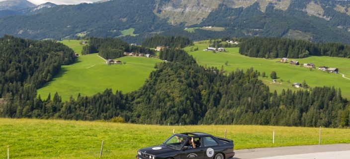 Ein schwarzes Auto fährt über eine Landstraße. Im Hintergrund Berge und Wäldchen.