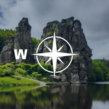 Kompassrose, das auf den Buchstaben "W" zeigt. Im Hintergrund ein zerklüfteter Feld an einem Fluss.