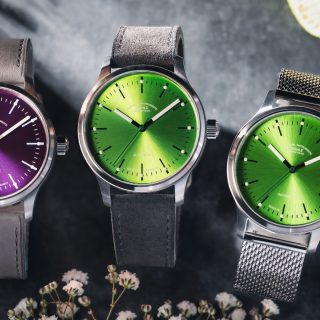 Drei Armbanduhren (Panova Violett am grauen Lederband, Panova Limettengrün am Veloursband und am Metallband) liegen nebeneinander auf einem grauen Tisch mit weißen Blüten.