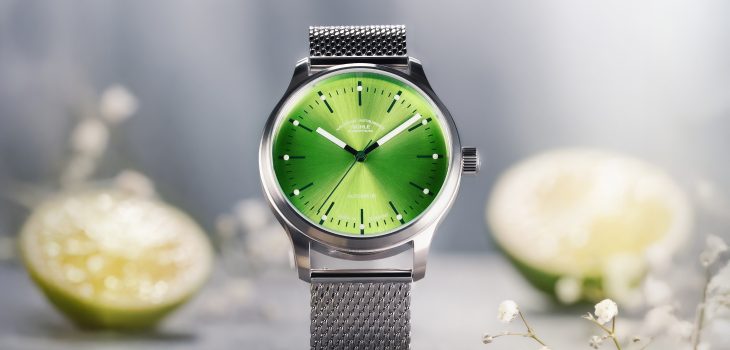 Frontalansicht der Armbanduhr Panova Limettengrün am Milanaiseband stehend auf einem hellen Tisch mit Limetten im Hintergrund.