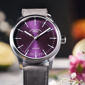 Frontalansicht der Panova Violett am grauen Lederband, stehend auf einem hellgrauen Tisch mit Blüten.