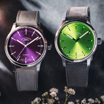 Zwei Armbanduhren (Panova Violett am grauen Lederband und Panova Limettengrün am grauen Veloursband) stehen nebeneinander auf einem grauen Tisch mit Blüten.