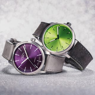 Zwei Armbanduhren (Panova Violett am grauen Lederband und Panova Limettengrün am grauen Veloursband) liegen seitlich übereinander auf einer hellgrauen Oberfläche.