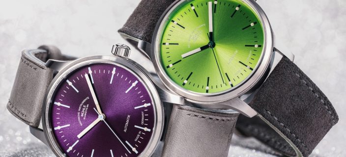 Zwei Armbanduhren (Panova Violett am grauen Lederband und Panova Limettengrün am grauen Veloursband) liegen seitlich übereinander auf einer hellgrauen Oberfläche.