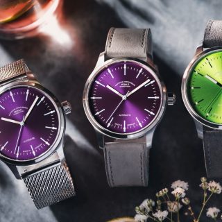 Drei Armbanduhren (Panova Violett mit Metallband, Panova Violett am grauen Lederband, Panova Limettengrün am grauen Veloursband) liegen nebeneinander auf einem grauen Tisch mit weißen Blüten.