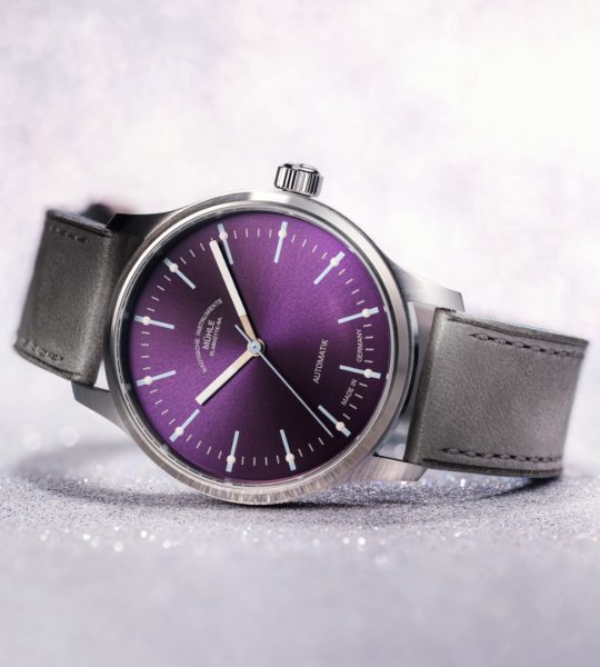 Armbanduhr Panova Violett am grauen Lederband liegt seitlich auf einem weißen Tisch.