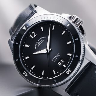Armbanduhr Sportivo Großdatum mit schwarzem Zifferblatt, schwarzer Lünette und silbernem Gehäuse am schwarzen Lederband lehnt leicht schräg an einer grauen Box.