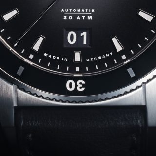 Detailbild der Armbanduhr Sportivo Großdatum mit schwarzem Zifferblatt, schwarzer Lünette und silbernem Gehäuse am schwarzen mit Fokus auf den Gehäuserand bei 6 Uhr.