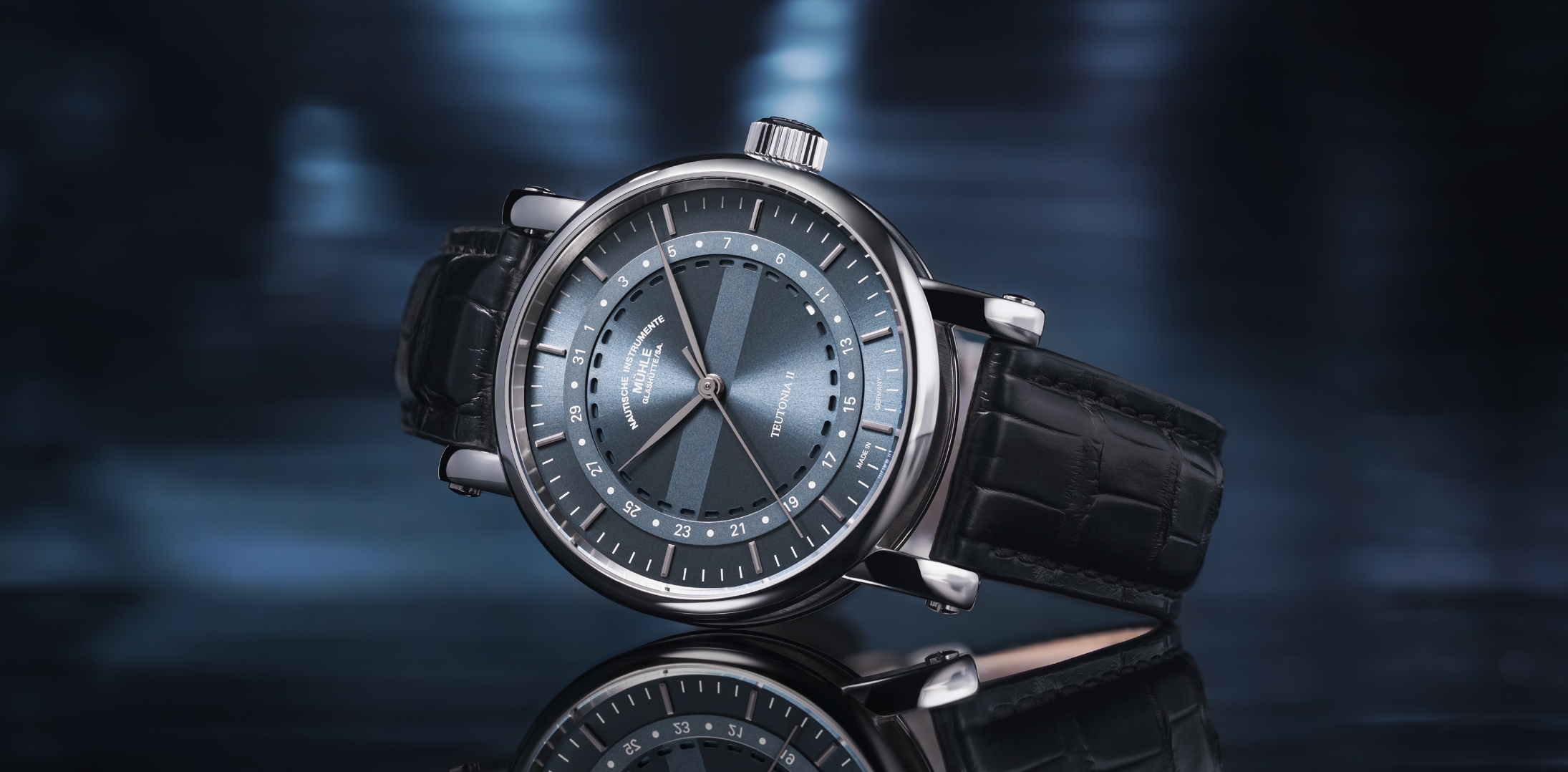 Teutonia II Ringdatum mit Krokolederband liegt seitlich auf spiegelnder Oberfläche.