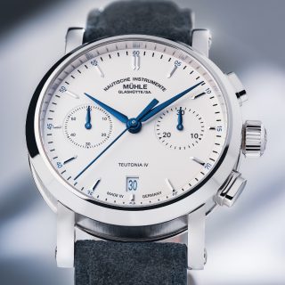 Frontalansicht der Armbanduhr Teutonia IV Chronograph mit weißem Zifferblatt im silbernen Gehäuse am blauen Velourslederband vor grauem Hintergrund.