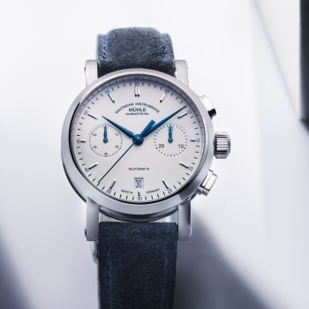 Teutonia IV Chronograph mit weißem Zifferblatt im silbernen Gehäuse am blauen Velourslederband steht auf einem weißen Untergrund.