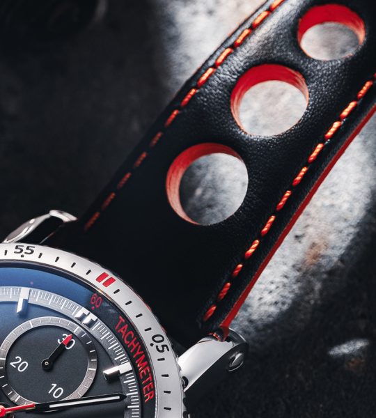Makroaufnahme eines schwarz-roten Lederbandes an einer Armbanduhr mit schwarzem Zifferblatt und silbernem Gehäuse.